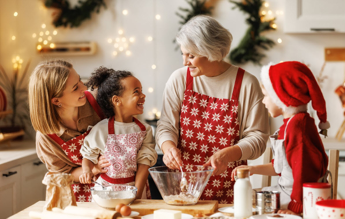 An older woman wearing a red apron has ingredients in a bowl while two young children and a woman smile and watch