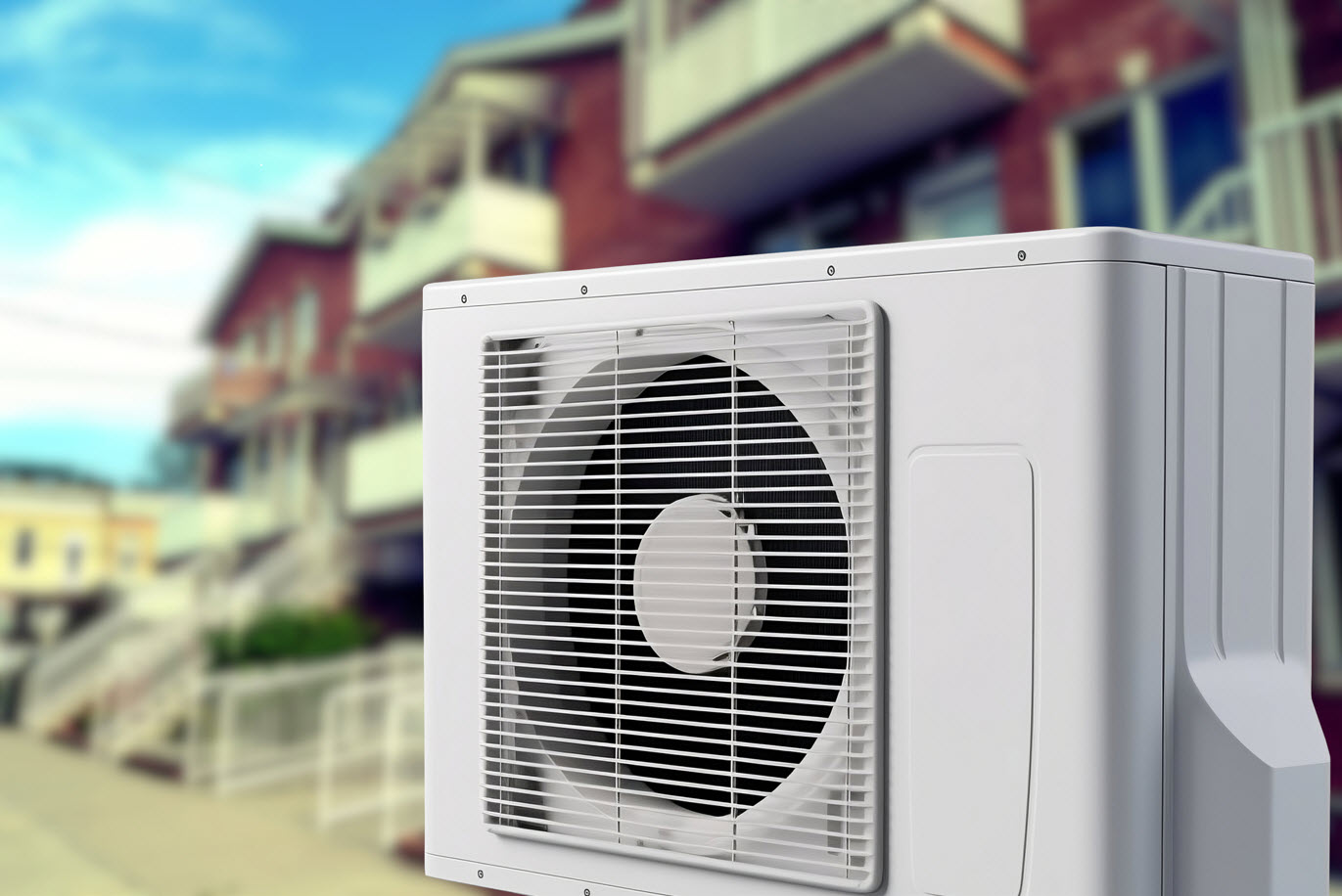 A heat pump is shown in close up with a backdrop of townhomes