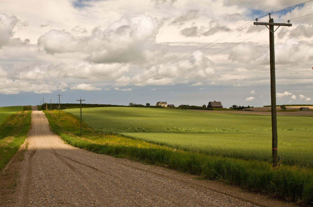 A rural dirt road with green fields, rolling clouds and farm homes in the distance. 