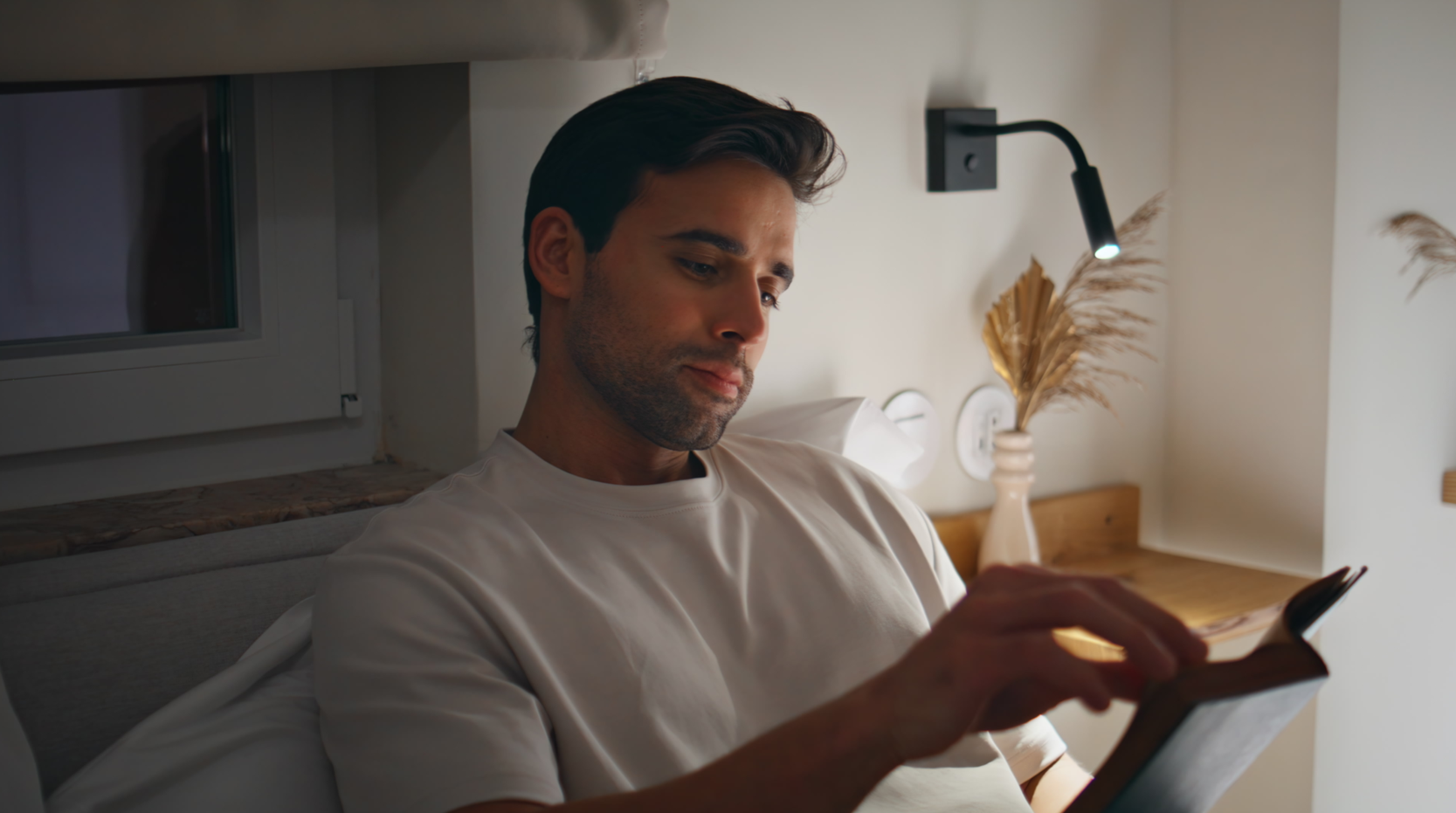 A man reads in bed next to a small wall lamp