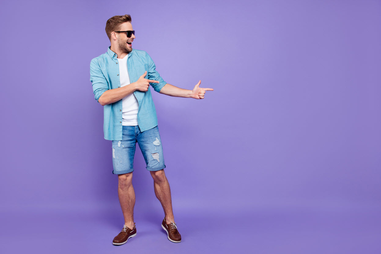 A young man in jean shorts poses with both hands pointing to the right