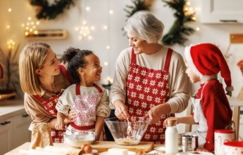 An older woman wearing a red apron has ingredients in a bowl while two young children and a woman smile and watch