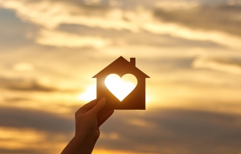 A person holds a cutout of a house with a heart-shape in the middle with the sun shining through