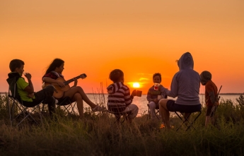 A family sit in camping chairs as the sun sets over a lake in the background