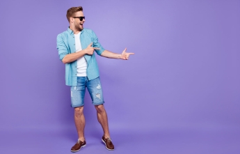 A young man in jean shorts poses with both hands pointing to the right