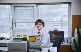 A man with a moustache and what appears to be a wig sits at an office desk