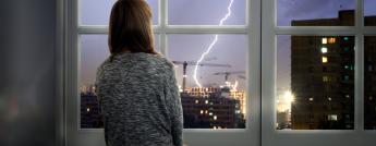 A woman looks out a window at a lightning storm