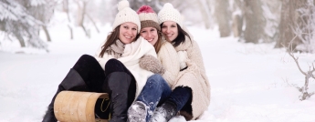 Three women dressed in hats and coats sit on a toboggan in a winter forest
