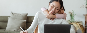 A woman works at her laptop while her daughter hugs her