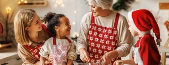 An older woman wearing a red apron has ingredients in a bowl while two young children and a woman smile and watch