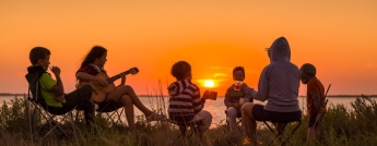 A family sit in camping chairs as the sun sets over a lake in the background