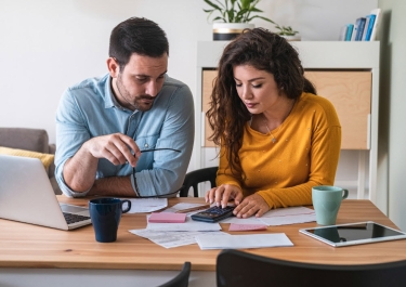 A couple calculates their bills over coffee at a kitchen table with a laptop and cacluclator